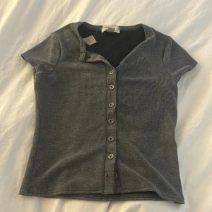 Grå ribbad topp med knappar - Säljer en grå ribbad topp från urban outfiters med korta ärmar och knappar hela vägen framtill. Enkel och stilren design som passar perfekt till jeans eller kjol. Toppen är i mjukt material och har en figurnära passform. helt oanvänd 
