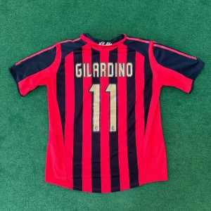AC Milan fotbollströja Gilardino 11 - Säljer en klassisk AC Milan fotbollströja med Gilardino 11 på ryggen. Tröjan är röd och svart med vertikala ränder, Adidas-logga och klubbmärke på bröstet samt sponsortryck. Kortärmad modell i lätt och ventilerande material.