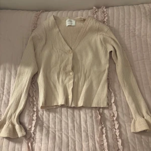 Beige ribbad kofta från Zara - Säljer en beige ribbad kofta från Zara med v-ringning och knappar framtill. Tröjan har långa ärmar och är croppad i modellen, perfekt att styla till olika outfits.