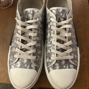 Gråvita sneakers från Dior - Säljer ett par gråvita Dior sneakers med tydligt monogrammönster och vita snören. Skorna har rund tå, vit gummisula och diskret Dior-logga på sidan. Perfekta för dig som gillar lyxiga detaljer och stilren design.
