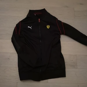 Svart Ferrari jacka från Puma - Säljer en svart track jacket från Puma i samarbete med Ferrari. Jackan har röda detaljer längs axlarna, dragkedja framtill och Ferrari-logga på bröstet. Perfekt för dig som gillar sportig stil och motorsport.