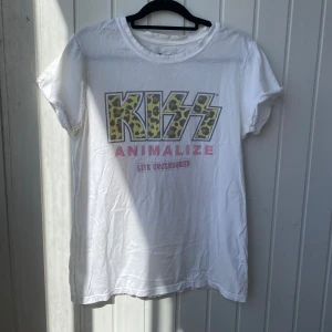 Vit KISS Animalize t-shirt från Recycled Karma - Säljer en vit KISS Animalize t-shirt från Recycled Karma. T-shirten har ett leopardmönstrat KISS-tryck och texten 'Animalize Live Uncensored' i rosa. Den är kortärmad och tillverkad i 100% bomull. Har ett litet hål vid armen men inget som syns vid användning ( se bild 3)