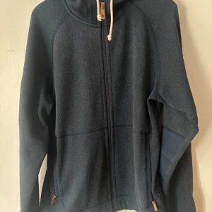 Mörkblå hoodie från Fjällräven - Säljer en mörkblå hoodie från Fjällräven i storlek M. Tröjan har dragkedja framtill, huva med snörning och snygga läderdetaljer vid snöret. Perfekt för kyliga dagar och outdoor-aktiviteter.