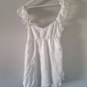 Vit volangklänning med knyt i ryggen - Söt vit klänning med volangdetaljer på axelbanden och v-ringad hals. Knytning i ryggen för extra detalj. Perfekt för sommarens alla tillfällen. Lätt och luftig modell med kort längd.