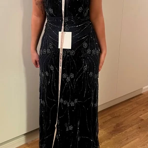 Mörkblå långklänning med glittriga detaljer - Säljer en elegant mörkblå långklänning med glittriga paljettdetaljer och blommigt mönster. Klänningen har v-ringning, transparent cape-liknande överdel och öppen rygg. Perfekt för bal eller fest. Figurnära passform och vackert fall.