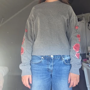 Grå sweatshirt med rosor - Säljer en grå sweatshirt med vackra röda rosor broderade längs ärmarna. Tröjan har en rund hals och ribbade muddar vid ärmslut och nederkant. Perfekt för en avslappnad stil.