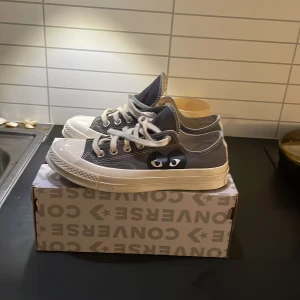 Converse x Comme des Garçons Play sneakers grå - Säljer ett par grå Converse x Comme des Garçons Play sneakers med det ikoniska röda hjärtat på sidan. Skorna har vita snören, vit tå och sula samt klassisk låg modell. Perfekta för dig som vill ha något unikt och stilrent.
