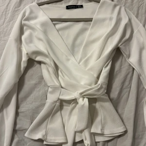 Vit omlottopp med knyt från Boohoo - Säljer en stilren vit omlottopp från Boohoo med lång ärm och knyt i midjan. Toppen har v-ringad hals och markerad midja som ger en snygg siluett. Perfekt att styla med jeans eller kjol.
