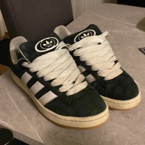 Svarta Adidas Campus sneakers - Säljer ett par klassiska svarta Adidas Campus sneakers med vita detaljer och snörning. Skorna har den ikoniska trefoil-loggan och tre ränder på sidan. Perfekta för en stilren look.