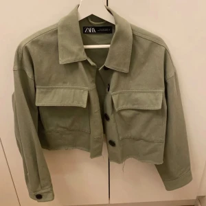 Olivgrön jacka från Zara - Säljer en olivgrön jacka från Zara med stora fickor och knappar framtill. Jackan har en croppad design och långa ärmar, perfekt för en trendig look. Passar bra till både vardag och speciella tillfällen.