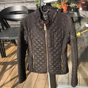 Svart quiltad jacka från Hollies - Säljer en svart quiltad jacka från Hollies i storlek 34. Jackan har guldiga dragkedjor, ståkrage och ett rutigt innerfoder. Snygg passform och perfekt för övergångssäsonger. Klassisk och stilren design.