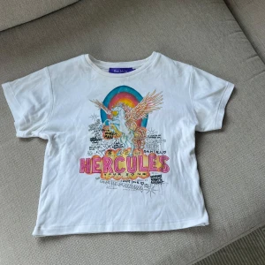 Vit t-shirt från Réalisation - Vit t-shirt med färgglatt Pegasus- och regnbågstryck samt texten 'Hercules'. Den kommer från realisation par och köpt för cirka 860kr🌈