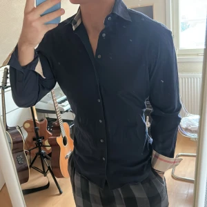 Burberry skjorta - Mörkblå svart skjorta från Burberry med klassiska rutiga detaljer på insidan av kragen och ärmsluten. Skjortan har en stilren design med knappar framtill och långa ärmar. Perfekt för en stilmedveten look.