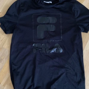Svart mesh t-shirt från Fila strl S - Säljer en svart t-shirt från Fila med stor logga framtill. T-shirten är gjord i meshmaterial som andas och har en sportig känsla. Perfekt för träning eller en avslappnad stil.