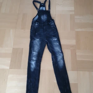 Svarta denim hängselbyxor från Rock Rebel - Säljer ett par svarta denim hängselbyxor från Rock Rebel med dödskalle-spännen. Byxorna har flera fickor och justerbara hängslen. De är för långa för mig som är 160 och passar antagligen någon som är runt 165-170