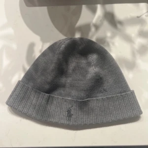 Grå stickad mössa från Polo Ralph Lauren - Snygg grå stickad mössa från Polo Ralph Lauren med diskret broderad logga framtill. Klassisk ribbad kant och mjukt material som håller dig varm under kyliga dagar. Har några hål i sig men inget som påverkar användningen 