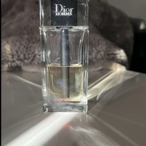 Dior Homme Sport Parfym - Säljer en elegant Dior Homme Sport parfym i en stilren glasflaska. Doften är fräsch och energisk, perfekt för den moderna mannen. Flaskan har en svart spraypump och tydlig märkning. En lyxig doft för alla tillfällen.