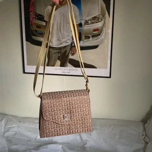 Beige axelväska med mönster - Säljer en beige axelväska med ett stilrent geometriskt mönster. Väskan har en justerbar axelrem i ljus färg och ett metallspänne framtill. Perfekt för att bära dina nödvändigheter med stil.