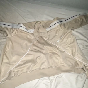 Beige jacka från Palm Angels - Säljer en snygg beige zip Äkta   från Palm Angels med vita och svarta ränder längs ärmarna. Zipen har tyvärr tappats bort men det går såklart att laga . Perfekt för en stilren look. Tyvärr så har den också hål i både inne fickorna. 