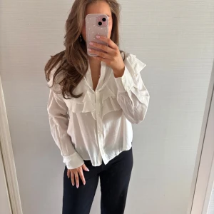 Vit blus med volangdetaljer - Säljer en stilren vit blus med volangdetaljer framtill. Blusen har långa ärmar och knappar längs framsidan. Använd enstaka gånger. Storlek L men jag har S/M i vanliga fall och ni ser hur den sitter på mig.