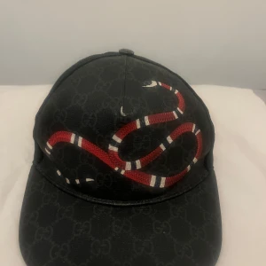 Svart keps med ormmotiv från Gucci - Snygg svart keps från Gucci med ett i ögonfallande rött ormmotiv. Kepsen har ett diskret mönster och justerbart band baktill med röd detalj. Perfekt för att ge din outfit en lyxig touch. Storlek S men Passar lika bra som storlek M