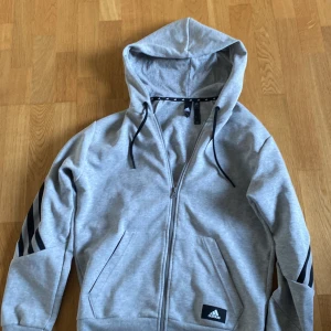 Grå hoodie från Adidas med svarta ränder - Säljer en grå hoodie från Adidas med dragkedja och huva. Tröjan har svarta ränder på ärmarna och två fickor framtill. Tillverkad i mjukt material och har Adidas-logga på ena fickan.