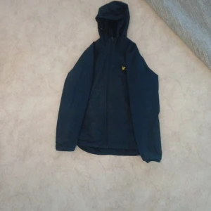 Mörkblå vindjacka med huva  - Säljer en mörkblå vindjacka från Adidas med huva och dragkedja framtill. Jackan har en liten gul Adidas-logga på bröstet och är perfekt för blåsiga dagar. Enkel och stilren design.