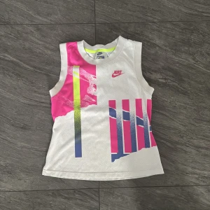 Vit och rosa ärmlös träningstopp från Nike - Nike Challenge Court träningstopp i vitt med färgglada rosa, blå och neongula detaljer. Ärmlös modell med sportig design och meshmaterial som andas. Perfekt för tennis eller annan träning.