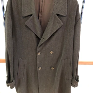 Vinterock Tiger - Stilren gråbrunsvart tunnfodrad vinterrock i ull med dubbelknäppning och bred krage. Jackan har långa ärmar, två snedställda fickor fram och ett bälte baktill. Perfekt för dig som vill ha en klassisk och tidlös look.