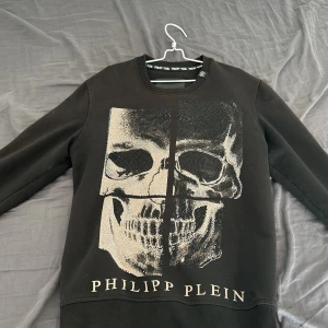 Svart sweatshirt med dödskalle från Philipp Plein - Cool svart sweatshirt från Philipp Plein med en stor dödskalle framtill dekorerad med glittrande stenar. Tröjan har rund halsringning och ribbade muddar. Märkeslogga på baksidan vid nacken. Perfekt statement-piece för dig som gillar edgy stil.