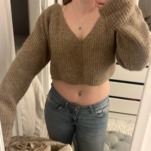Beige croppad stickad tröja med v-ringning - Supermjuk och croppad stickad tröja i beige med djup v-ringning både fram och bak. Tröjan har långa ärmar och en oversized passform, perfekt för lager-på-lager-stil.