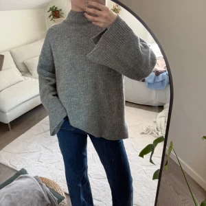 Grå stickad polotröja med vida ärmar - Mysig grå stickad polotröja med vida ärmar, har även en slits i ärmarna. Enkel att styla med jeans.