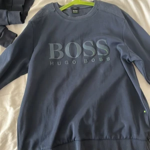 Blå sweatshirt från Hugo Boss - Säljer en stilren blå sweatshirt från Hugo Boss med deras logotyp tryckt på framsidan. Tröjan har långa ärmar och en rund halsringning. Perfekt för en avslappnad stil.