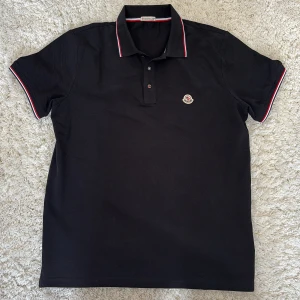 Svart pikétröja från Moncler - Snygg svart pikétröja från Moncler med klassisk krage och knappar. Tröjan har röda och vita detaljer på kragen och ärmsluten samt Monclers logga broderad på bröstet. Perfekt för en stilren look! Pikén är helt ny och bara provad, säljer den för att den var en aning stor. Hör av dig om du har några funderingar eller frågor om tröjan, kan gå ner i pris vid snabb affär!