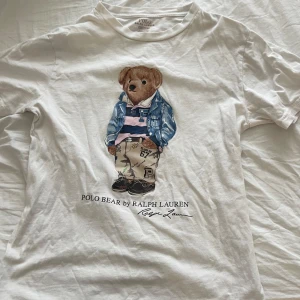 Vit t-shirt med Polo Bear från Ralph Lauren - Vit t-shirt från Ralph Lauren med ikoniska Polo Bear-trycket på bröstet. Björnen har på sig jeansjacka, randig piké och beiga byxor. Klassisk rund halsringning och mjukt bomullsmaterial.
