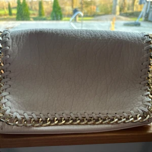 Beige axelväska med guldkedja från Tiamo - Säljer en stilren beige axelväska från Tiamo med grov guldkedja som detalj runt kanterna. Väskan har en dragkedja på baksidan och ett rymligt fack inuti. Perfekt för att lyfta din outfit med en trendig accessoar.