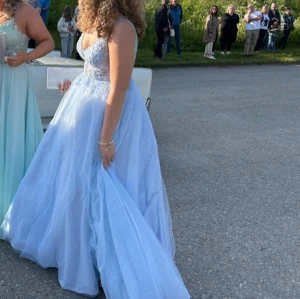 Ljusblå glittrig balklänning - Elegant ljusblå balklänning med glittrande detaljer och djup v-ringning. Klänningen har en lång, flödande kjol och är ärmlös, vilket ger en sofistikerad look. Perfekt för en speciell kväll.