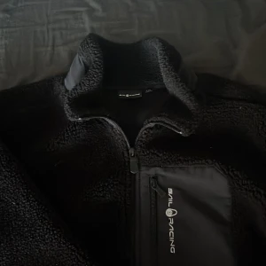 Svart fleecejacka från Sail Racing (HELST BYTEN) - Säljer en svart fleecejacka från Sail Racing med dragkedja och en bröstficka. Jackan har en hög krage och är perfekt för kyliga dagar. Den är både stilren och funktionell. Skulle helst vilja byta mot någon typ av kofta/ hoodie i storlek XS/170