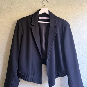 Kort blazer/kavaj i svart  - En kortare kavaj/blazer i svart. Stängs med en knapp i svart i mitten på magen. Långa armar med knappar längs underarmarna. Axelkuddar förekommer, men är diskreta! Modellen är kortare baktill än framtill. Sparsamt använd, inga tecken på slitage. Mått: 50cm lång i ryggen, 70 cm lång framtill och 45 cm axel till axel 🐝