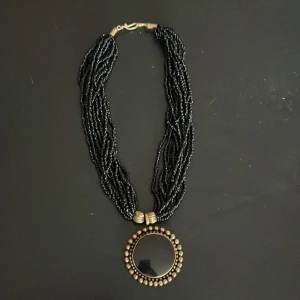 Svart halsband med stor rund medaljong - Unikt halsband med flera rader svarta pärlor och en stor rund medaljong i svart och antikguld. Medaljongen har en ram av små guldfärgade kulor som ger ett coolt statement-utseende. Perfekt för dig som gillar att sticka ut med accessoarer.