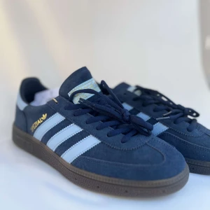 Adidas Handball Spezial marinblå sneakers - Adidas Handball Spezial sneakers i marinblå mocka med klassiska vita ränder och guldfärgad Spezial-logga på sidan. Snygg retrostil med svart sula och snörning. Perfekta för dig som gillar streetwear och tidlös design.