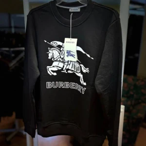 Svart Burberry sweatshirt  - Svart sweatshirt från Burberry i 100% bomull med stort vitt tryck av riddare och häst på bröstet samt Burberry-logga. Klassisk rund halsringning och långa ärmar. Perfekt för dig som vill ha en stilren och lyxig streetwear-look.finns i fler storlekar 