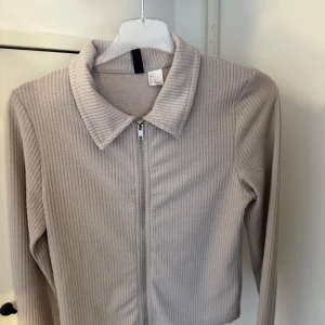 Beige ribbad kofta med dragkedja - Säljer en beige ribbad kofta från H&M med dragkedja framtill och klassisk krage. Långärmad och i mjukt material som passar perfekt till lager-på-lager. Enkel och stilren design som är lätt att matcha.