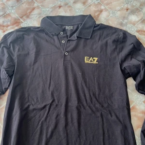 Svart pikétröja EA7 Emporio Armani - Svart pikétröja från EA7 Emporio Armani med klassisk krage och tre knappar framtill. Snygg gul logga på bröstet och diskreta gula ränder på ryggen. Tillverkad i mjuk bomull, perfekt för en clean och stilren look.