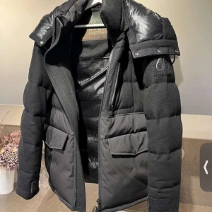 Svart pufferjacka Moose Knuckles - Säljer en svart pufferjacka från Moose Knuckles med coola detaljer och stor huva. Jackan har stora fickor framtill, dragkedja och är tillverkad i en mix av ull och syntetmaterial. Perfekt för kalla dagar och har en riktigt modern vibe. kvitto finns också.. Kom DM om ni har funderingar.