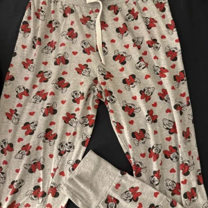 Disney mjukisbyxor med Minnie Mouse - Grå mjukisbyxor från Disney med gulligt Minnie Mouse-tryck och röda hjärtan över hela byxan. Byxorna har dragsko i midjan och muddar vid bensluten. Perfekta för chill dagar hemma eller sleepovers.