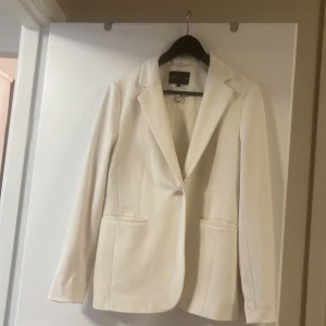 Vit kavaj från Stockholm LM (MQ) - Stilren vit kavaj från MQ med klassisk krage och en knapp framtill. Kavajen har två fickor och figursydd passform som ger en snygg siluett. Perfekt för dig som vill ha en fräsch och clean look.