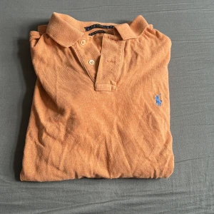 Orange pikétröja från Polo Ralph Lauren - Snygg orange pikétröja från Polo Ralph Lauren med klassisk krage, två knappar och blå broderad logga på bröstet. Modellen är custom slim fit och tillverkad i mjuk bomull. Perfekt för dig som gillar en stilren och avslappnad look. Hör gärna av er vid minsta lilla fråga 