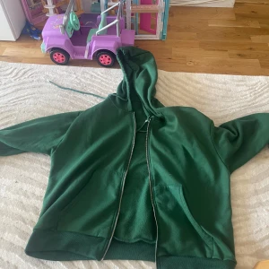 Grön hoodie med dragkedja - Mörkgrön hoodie med huva och dragkedja framtill. Tröjan har känguruficka och snörning i huvan. Perfekt för chill dagar eller när du vill ha en avslappnad stil.