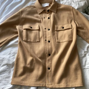 Beige overshirt från Selected Homme - Stilren beige overshirt från Selected Homme med klassisk krage, två stora bröstfickor med lock och svarta knappar framtill. Overshirten har lång ärm och är tillverkad i ett mjukt material som känns skönt mot huden. Perfekt att slänga över en t-shirt för en chill look.